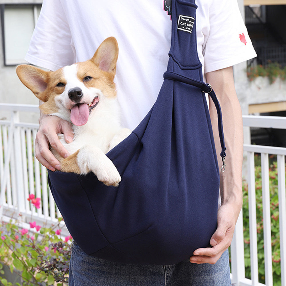Corgi dog in a navy blue sling held by a person outdoors.