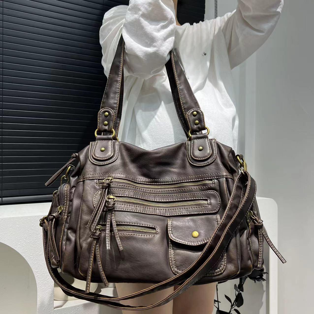 Brown leather handbag with multiple zippers held by a person wearing a white shirt.