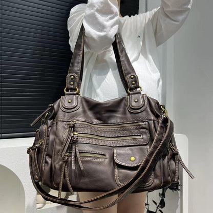 Brown leather handbag with multiple zippers held by a person wearing a white shirt.