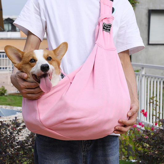Person holding a pink dog sling with a corgi inside, outdoors.