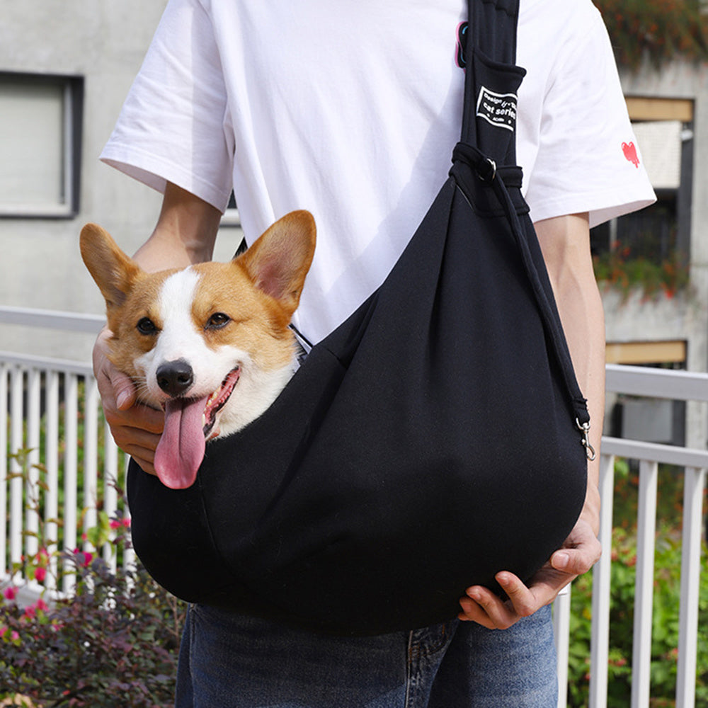 Person holding a black pet sling with a corgi dog inside, outdoors.